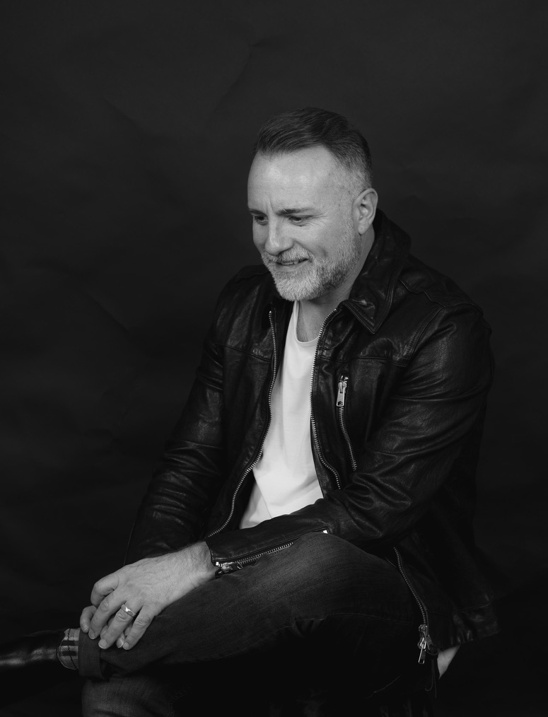 Black-and-white portrait of Mark Stevens seated in a leather jacket